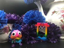 Load image into Gallery viewer, image is a rainbow (pink, red, orange, yellow, green, turquoise, blue, purple) guillotine earring with a silver blade dangling from the top. there is a small jump ring visible at the bottom of the guillotine.   background of image has blue and purple silk flowers (carnations) and a white floral-patterned teapot and teacup. next to the earring is a small rainbow penis with googly eyes. 
