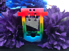 Load image into Gallery viewer, image is a rainbow (pink, red, orange, yellow, green, turquoise, blue, purple) guillotine earring with a silver blade dangling from the top. there is a small jump ring visible at the bottom of the guillotine. There are white specks of paint visible through the different colors of the rainbow.  background of image has blue and purple silk flowers (carnations). a white, floral-patterned teapot is barely visible in the back right corner.
