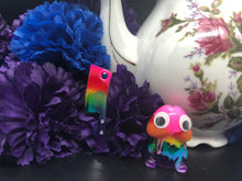 Load image into Gallery viewer, image is a rainbow(pink, red, orange, yellow, green, turquoise, blue, purple) butcher knife earring.  background of image has blue and purple silk flowers (carnations) and a white floral-patterned teapot and teacup. next to the earring is a small rainbow penis with googly eyes. 
