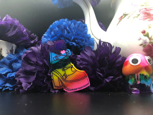 image is a reverse rainbow(pink, red, orange, yellow, green, turquoise, blue, purple from the bottom up) heeled boot with a pink heart and laces engraved with purple paint.   background of image has blue and purple silk flowers (carnations) and a white floral-patterned teapot and teacup. next to the earring is a small rainbow penis with googly eyes. 