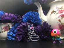 Load image into Gallery viewer, image is a dark purple, sparkly, heeled boot with a pink heart and laces engraved with white paint.   background of image has blue and purple silk flowers (carnations) and a white floral-patterned teapot and teacup. next to the earring is a small rainbow penis with googly eyes. 
