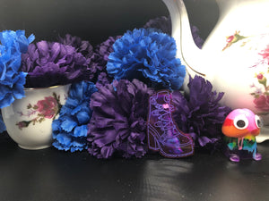 image is a dark purple heeled boot with a pink heart and laces engraved with purple paint.   background of image has blue and purple silk flowers (carnations) and a white floral-patterned teapot and teacup. next to the earring is a small rainbow penis with googly eyes. 