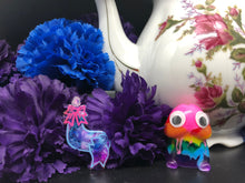 Load image into Gallery viewer, image is a clear furry tail plug earring with swirls of pink, purple, and blue. The engravings are done in a light blue and pink paint.  background of image has blue and purple silk flowers (carnations) and a white floral-patterned teapot and teacup. next to the earring is a small rainbow penis with googly eyes. 
