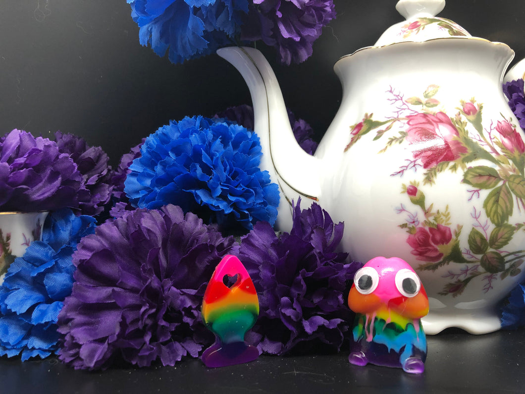 image is a rainbow(pink, red, orange, yellow, green, turquoise, blue, purple) plug earring.  background of image has blue and purple silk flowers (carnations) and a white floral-patterned teapot and teacup. next to the earring is a small rainbow penis with googly eyes. 