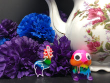 Load image into Gallery viewer, image is a rainbow(pink, red, orange, yellow, green, turquoise, blue, purple) furry tail plug earring with white paint engraving.  background of image has blue and purple silk flowers (carnations) and a white floral-patterned teapot and teacup. next to the earring is a small rainbow penis with googly eyes. 
