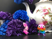 Load image into Gallery viewer, image is a pink and white swirled furry tail plug earring with white paint engraving.  background of image has blue and purple silk flowers (carnations) and a white floral-patterned teapot and teacup. next to the earring is a small rainbow penis with googly eyes. 

