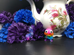 image is a pink, blue, and white swirled furry tail plug earring with white paint engraving.  background of image has blue and purple silk flowers (carnations) and a white floral-patterned teapot and teacup. next to the earring is a small rainbow penis with googly eyes. 