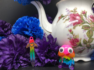 image is a  rainbow(pink, red, orange, yellow, green, turquoise, blue, purple) short sword with black paint engraving.  background of image has blue and purple silk flowers (carnations) and a white floral-patterned teapot and teacup. next to the earring is a small rainbow penis with googly eyes. 