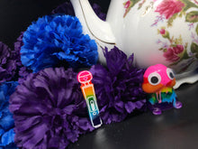 Load image into Gallery viewer, image is a rainbow(pink, red, orange, yellow, green, turquoise, blue, purple) magic wand vibrator with white engraving paint.  background of image has blue and purple silk flowers (carnations) and a white floral-patterned teapot and teacup. next to the earring is a small rainbow penis with googly eyes. 
