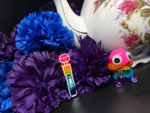 image is a rainbow(pink, red, orange, yellow, green, turquoise, blue, purple) magic wand vibrator with white engraving paint.  background of image has blue and purple silk flowers (carnations) and a white floral-patterned teapot and teacup. next to the earring is a small rainbow penis with googly eyes. 
