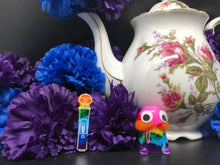 Load image into Gallery viewer, image is a rainbow(pink, red, orange, yellow, green, turquoise, blue, purple) magic wand vibrator with white engraving paint.  background of image has blue and purple silk flowers (carnations) and a white floral-patterned teapot and teacup. next to the earring is a small rainbow penis with googly eyes. 
