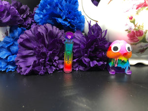 image is a reverse rainbow(pink, red, orange, yellow, green, turquoise, blue, purple from the bottom up) magic wand vibrator with black engraving paint and a small pink heart.  background of image has blue and purple silk flowers (carnations) and a white floral-patterned teapot and teacup. next to the earring is a small rainbow penis with googly eyes. 
