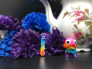 image is a reverse rainbow(pink, red, orange, yellow, green, turquoise, blue, purple from the bottom up) magic wand vibrator with white paint engraving.  background of image has blue and purple silk flowers (carnations) and a white floral-patterned teapot and teacup. next to the earring is a small rainbow penis with googly eyes. 