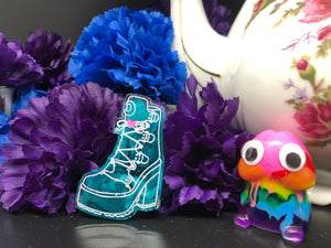 image is a teal high-heeled boot earring, with a pink heart and white paint engraving.  background of image has blue and purple silk flowers (carnations) and a white floral-patterned teapot and teacup. next to the earring is a small rainbow penis with googly eyes. 