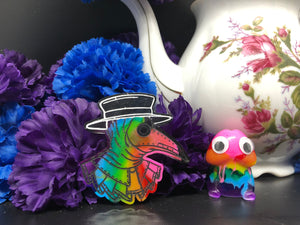 image is a image is a rainbow(pink, red, orange, yellow, green, turquoise, blue, purple) plague doctor with a black hat.  background of image has blue and purple silk flowers (carnations) and a white floral-patterned teapot and teacup. next to the earring is a small rainbow penis with googly eyes. 