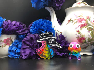image is a image is a rainbow(pink, red, orange, yellow, green, turquoise, blue, purple) plague doctor with a black hat.  background of image has blue and purple silk flowers (carnations) and a white floral-patterned teapot and teacup. next to the earring is a small rainbow penis with googly eyes. 