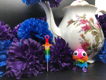 Load image into Gallery viewer, image is a image is a rainbow(pink, red, orange, yellow, green, turquoise, blue, purple) long sword earring with white paint engraving.  background of image has blue and purple silk flowers (carnations) and a white floral-patterned teapot and teacup. next to the earring is a small rainbow penis with googly eyes. 

