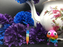 Load image into Gallery viewer, image is a reverse rainbow(pink, red, orange, yellow, green, turquoise, blue, purple from the bottom up) long sword earring with black paint engraving.  background of image has blue and purple silk flowers (carnations) and a white floral-patterned teapot and teacup. next to the earring is a small rainbow penis with googly eyes. 
