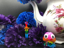 Load image into Gallery viewer, image is a rainbow(pink, red, orange, yellow, green, turquoise, blue, purple) long sword earring with black paint engraving. background of image has blue and purple silk flowers (carnations) and a white floral-patterned teapot and teacup. next to the earring is a small rainbow penis with googly eyes.
