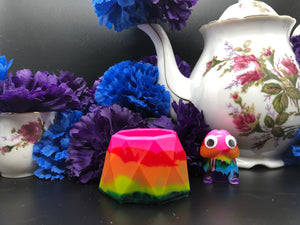 image is a rainbow(pink, red, orange, yellow, green, turquoise, blue, purple) trinket dish.   background of image has blue and purple silk flowers (carnations) and a white floral-patterned teapot and teacup. next to the earring is a small rainbow penis with googly eyes.