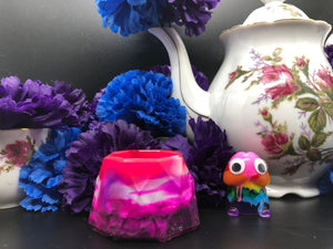 image is a pink, white, and purple layered and swirled trinket dish.  background of image has blue and purple silk flowers (carnations) and a white floral-patterned teapot and teacup. next to the earring is a small rainbow penis with googly eyes.