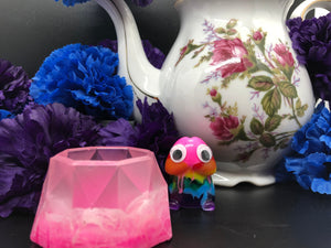 image is a light pink and white swirled trinket dish.   background of image has blue and purple silk flowers (carnations) and a white floral-patterned teapot and teacup. next to the earring is a small rainbow penis with googly eyes.