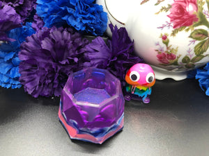 image is a small trinket dish with purple, blue, white, and pink swirls.  background of image has blue and purple silk flowers (carnations) and a white floral-patterned teapot and teacup. next to the earring is a small rainbow penis with googly eyes.