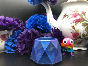 image is a blue/purple colorshift trinket dish.  background of image has blue and purple silk flowers (carnations) and a white floral-patterned teapot and teacup. next to the earring is a small rainbow penis with googly eyes.