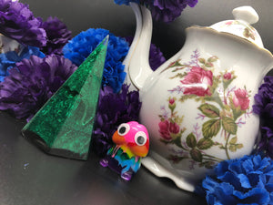 image is a small green, sparkly ring holder obelisk  background of image has blue and purple silk flowers (carnations) and a white floral-patterned teapot and teacup. next to the earring is a small rainbow penis with googly eyes.