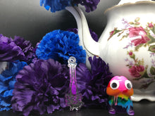 Load image into Gallery viewer, image is a dark purple to white ombre baseball bat earring with black paint engraving.  background of image has blue and purple silk flowers (carnations) and a white floral-patterned teapot and teacup. next to the earring is a small rainbow penis with googly eyes.
