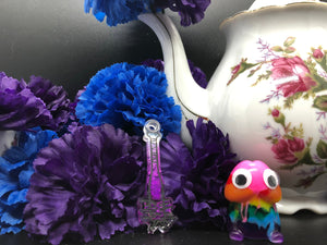 image is a dark purple to white ombre baseball bat earring with black paint engraving.  background of image has blue and purple silk flowers (carnations) and a white floral-patterned teapot and teacup. next to the earring is a small rainbow penis with googly eyes.