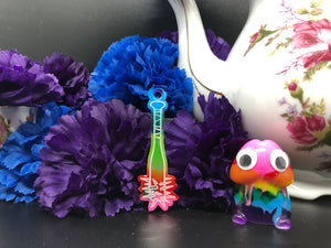 image is a rainbow(pink, red, orange, yellow, green, turquoise, blue, purple) baseball bat with white paint engraving.  background of image has blue and purple silk flowers (carnations) and a white floral-patterned teapot and teacup. next to the earring is a small rainbow penis with googly eyes.