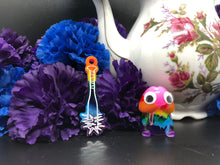Load image into Gallery viewer, image is a rainbow(pink, red, orange, yellow, green, turquoise, blue, purple) baseball bat with white paint engraving.  background of image has blue and purple silk flowers (carnations) and a white floral-patterned teapot and teacup. next to the earring is a small rainbow penis with googly eyes.

