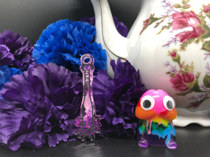 image is a dark purple to white ombre baseball bat earring with black paint engraving.  background of image has blue and purple silk flowers (carnations) and a white floral-patterned teapot and teacup. next to the earring is a small rainbow penis with googly eyes.