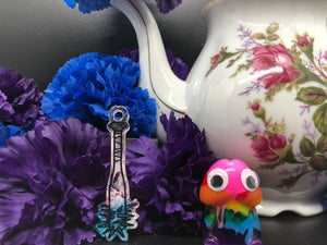 image is a blue, pink, and white baseball bat earring with black paint engraving.  background of image has blue and purple silk flowers (carnations) and a white floral-patterned teapot and teacup. next to the earring is a small rainbow penis with googly eyes.