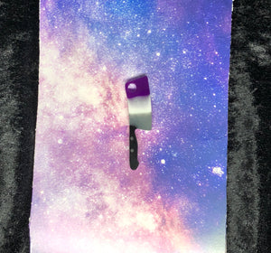 A kitchen cleaver earring with Ace Pride colors, purple white and grey on the blade and black on the handle. The background is a pink-and-blue nebula on black crushed velvet.