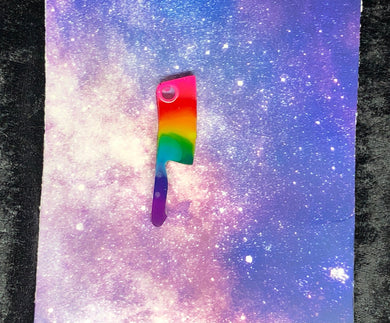 A kitchen cleaver earring with rainbow colors (pink, red, orange, yellow, green, teal, blue, and purple) against a pink-and-blue nebula and black crushed velvet background