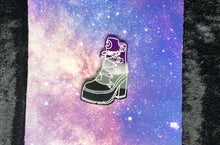 Load image into Gallery viewer, A broomrider boot earring with ace pride colors (black, grey, white, purple) and bright white highlights, against a pink-and-blue nebula and black crushed velvet background.
